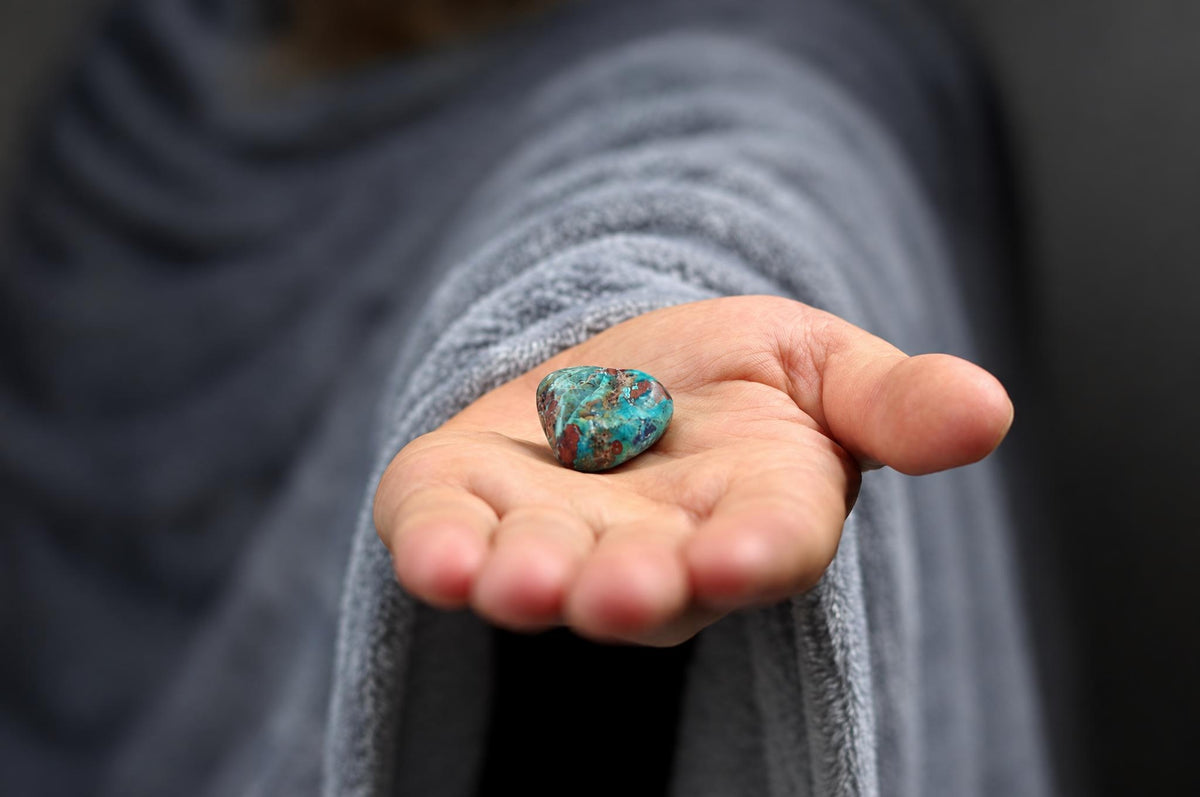 A hand holds a polished, heart-shaped stone with blue and reddish-brown patterns on a dark surface.