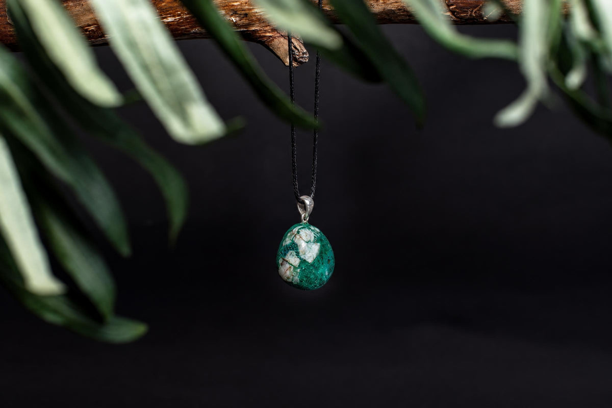 A green and white patterned pendant hangs from a black cord, suspended by a silver bail against a dark background with blurred leaves.