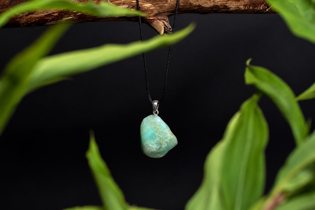 An irregular green chrysoprase pendant hangs from a black cord beneath a leafy branch against a dark background.