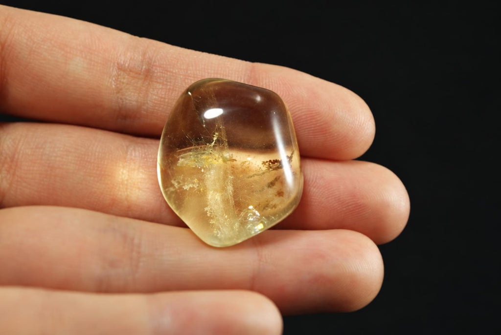 A polished, translucent stone with yellow and brown tones held in a hand against a dark surface.