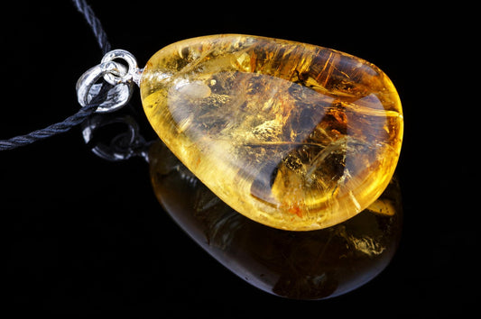 A polished, teardrop-shaped citrine pendant with golden-yellow tones and internal inclusions hangs from a silver bail on a dark cord against a black reflective surface.