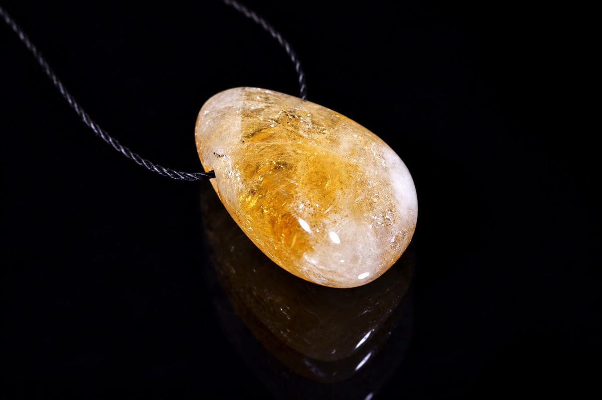 A polished citrine pendant with golden-yellow and white tones hangs on a dark cord against a black reflective surface.