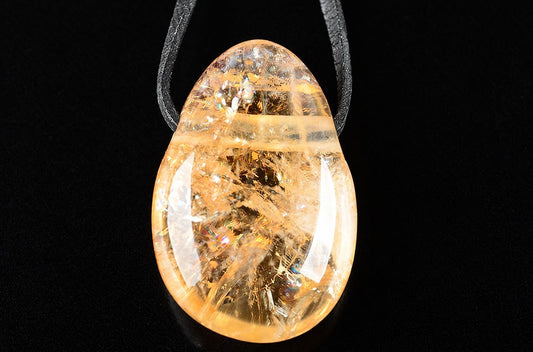 A teardrop-shaped pendant with a clear, yellow-orange stone containing golden inclusions, suspended on a dark cord against a black background.