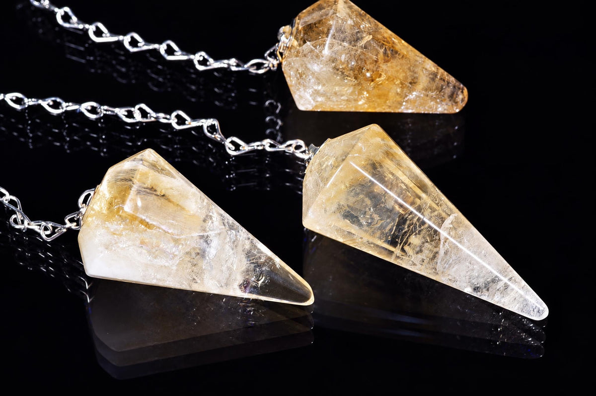 Three citrine pendulums with silver-toned chains rest on a dark, reflective surface.