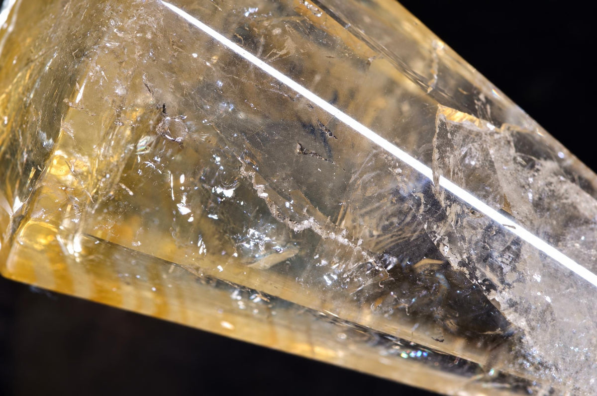 A close-up of a yellow citrine crystal point with visible internal fractures and inclusions against a dark background.