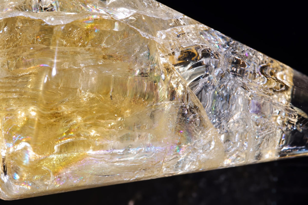 A close-up of a translucent, golden-yellow crystal point with internal inclusions and rainbow iridescence against a dark background.