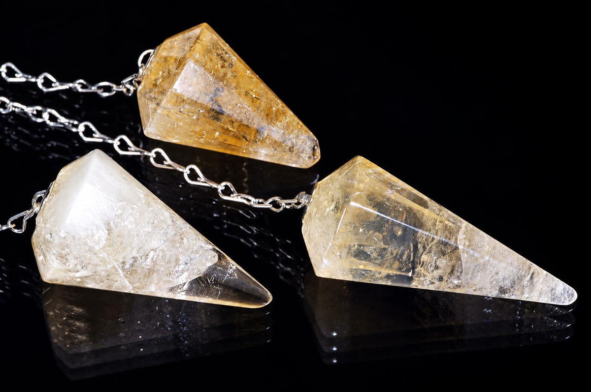 Three silver-toned chains hold clear, yellow citrine pendulums shaped like pointed crystals on a dark reflective surface.