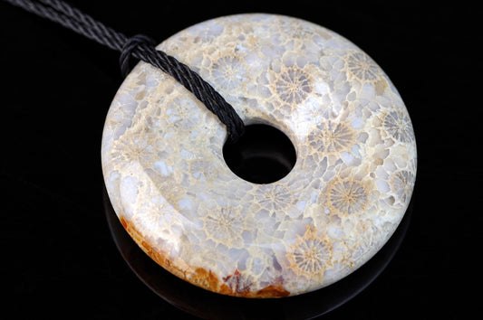 A donut-shaped pendant with a black cord, featuring a fossilized coral pattern in beige and white on a dark surface.