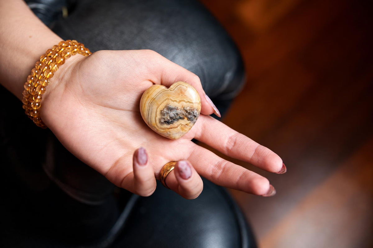 A hand holds a polished, heart-shaped piece of crazy lace agate with yellow and grey bands on a dark surface.