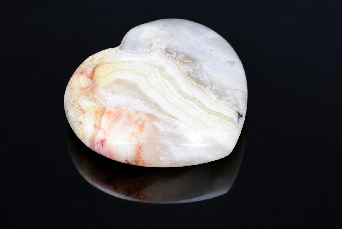 A polished, heart-shaped stone with white, yellow and pink banding on a dark reflective surface.