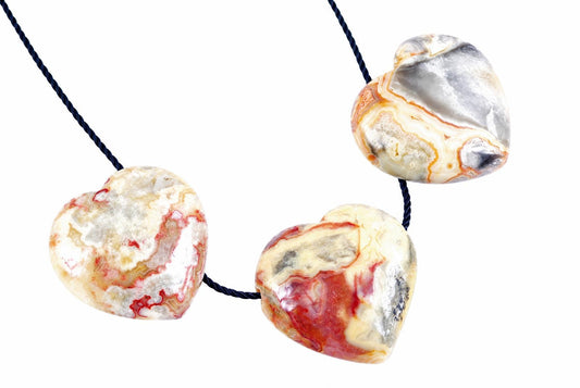 Three heart-shaped pendants made of multicoloured agate with red, white and grey patterns on a dark cord against a pale surface.