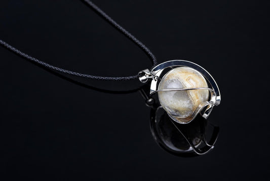 A silver-toned wire pendant holds a round, banded agate stone on a black cord against a dark surface.