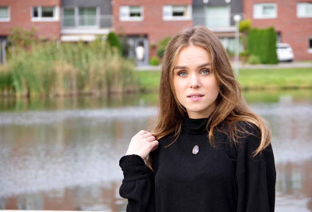 A woman wears a black turtleneck sweater with a silver-toned, oval-shaped pendant necklace against a blurred water and brick building background.