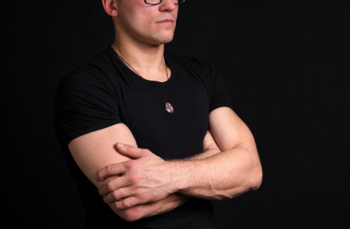 A man in a black t-shirt wears a pendant with a dark, textured oval stone on a thin cord against a dark background.