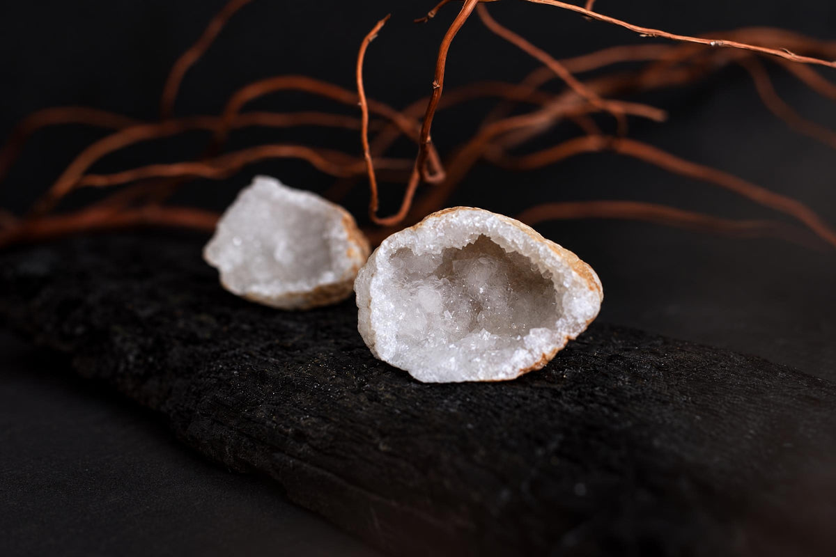 An open geode pair with white crystalline interiors rests on a dark textured surface with bare branches behind it.
