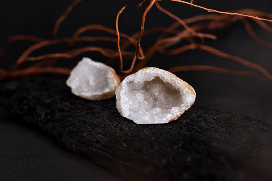An open geode pair with white crystalline interiors rests on a dark textured surface with bare branches behind it.