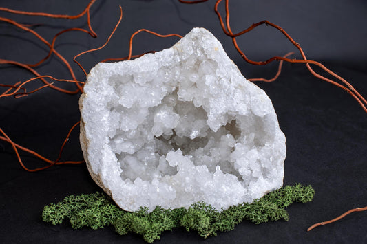A large, open white crystal geode with a rough outer shell sits on green moss against a dark surface.