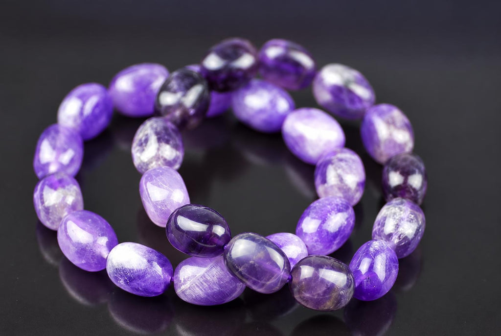 Overlapping loops of polished oval purple beads with pale inclusions and glossy highlights on a dark reflective surface, viewed at an angle with the rear beads softly out of focus.