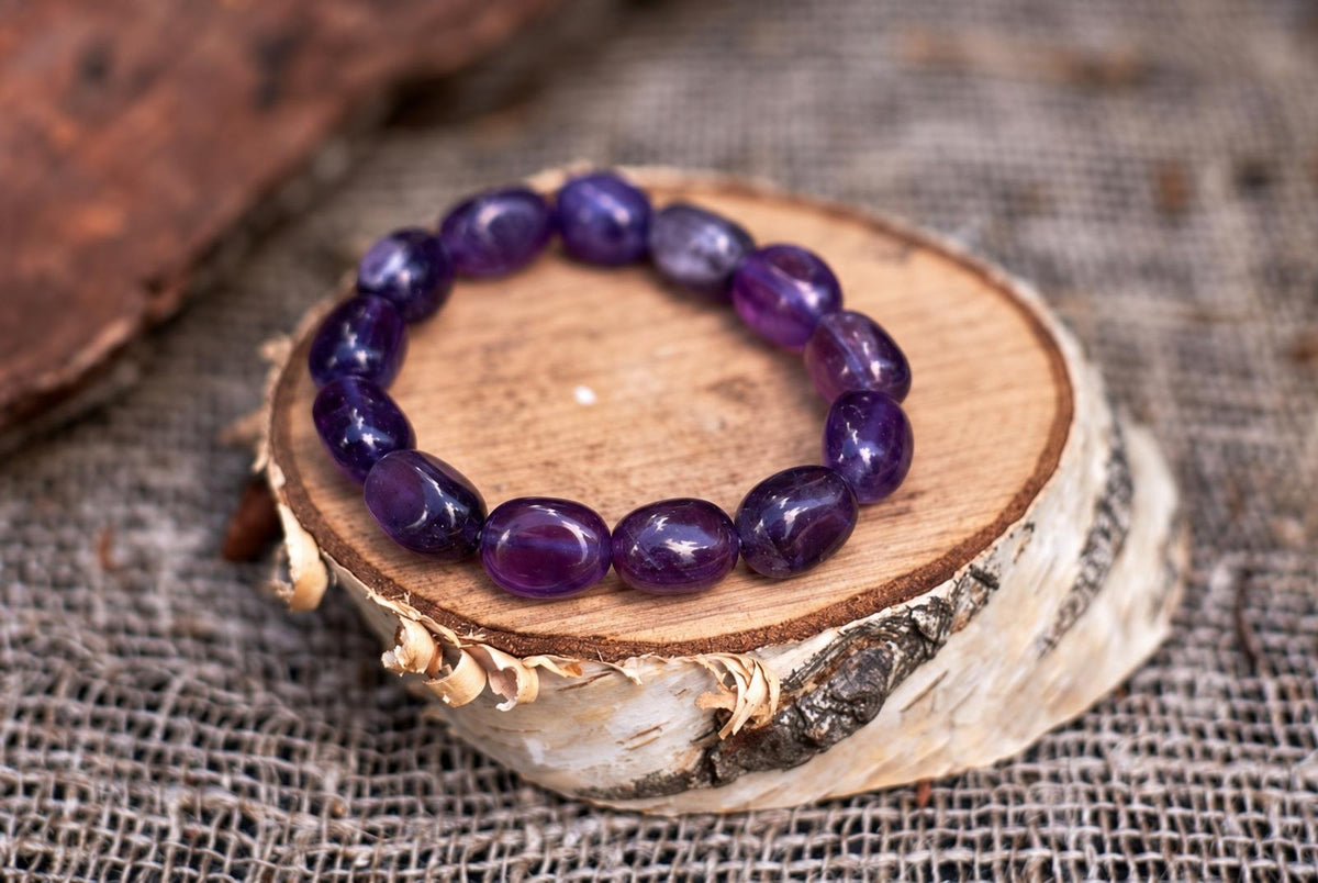 Glossy purple beads form a bracelet on a round, bark-edged wood slice resting on coarse woven fabric, viewed at an angle with a softly blurred background.