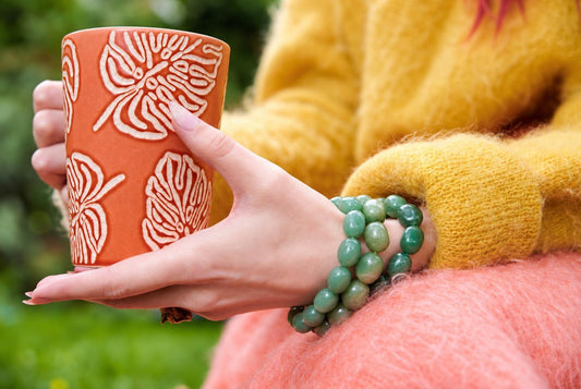 Close-up of green oval bead strands around a wrist beside a yellow knitted sleeve, with hands holding an orange cup patterned with white leaves against a blurred green outdoor background.