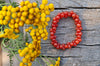 Glossy orange-red beaded bracelet arranged in an oval on weathered grey wood, beside softly blurred yellow flowers and green stems, viewed from above.