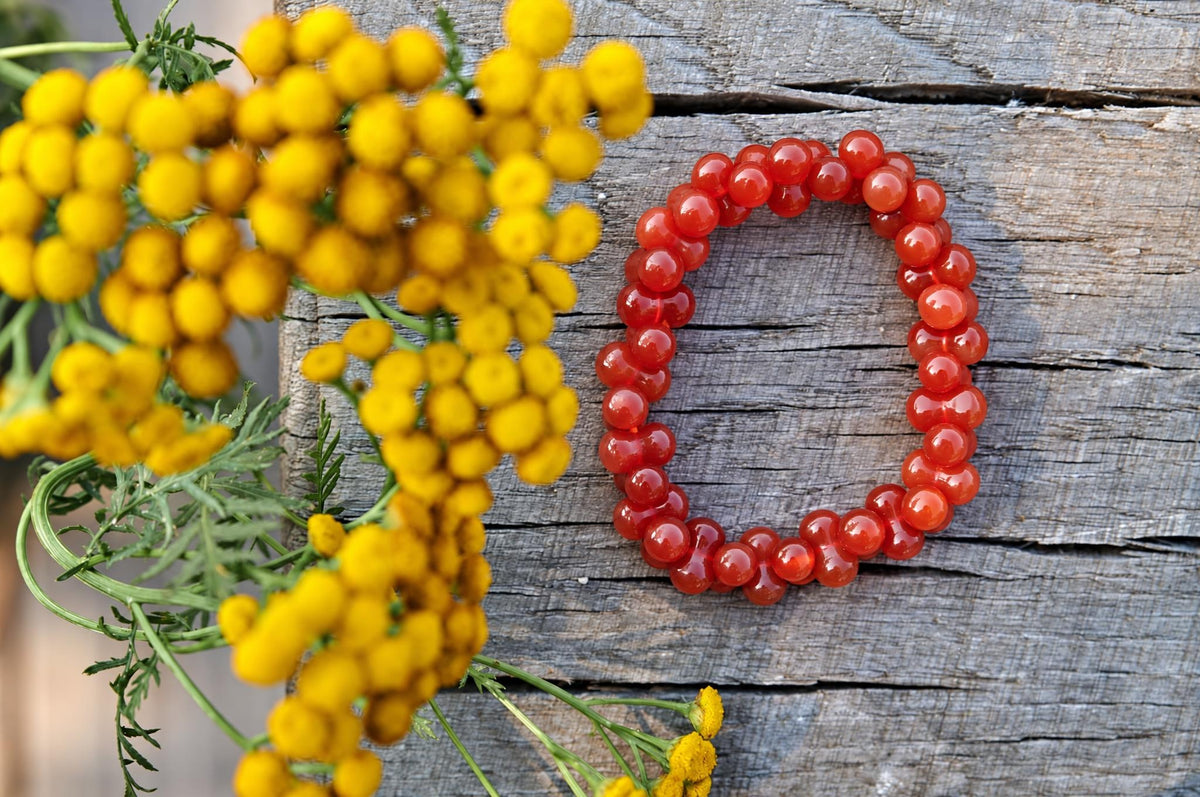 Glossy orange-red beaded bracelet arranged in an oval on weathered grey wood, beside softly blurred yellow flowers and green stems, viewed from above.