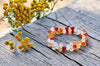 Bracelet of glossy round red, orange and milky translucent beads arranged in a loop on weathered wooden planks, beside small yellow flowers, viewed at an angle.