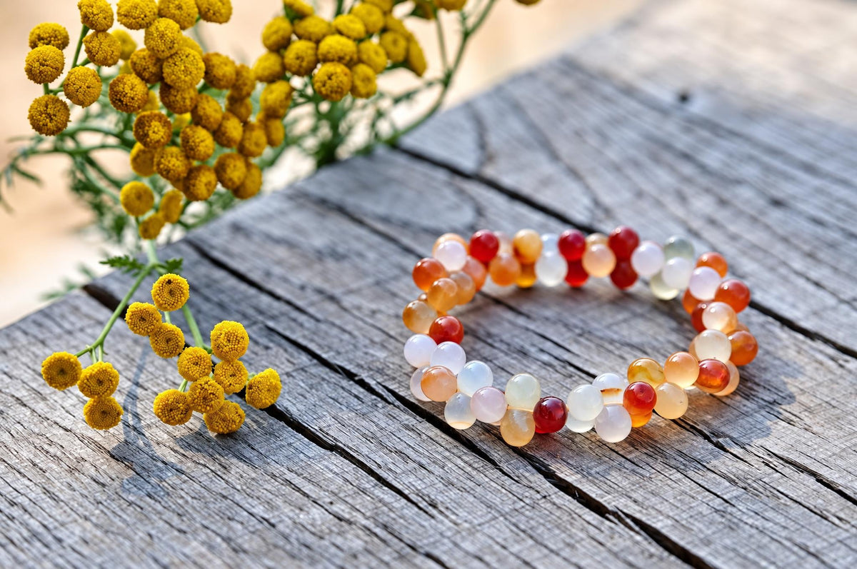 Bracelet of glossy round red, orange and milky translucent beads arranged in a loop on weathered wooden planks, beside small yellow flowers, viewed at an angle.