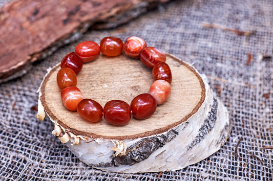 Glossy rounded red-orange bead bracelet with pale bands and a peach-coloured bead, arranged in a circle on a bark-edged wood slice over coarse woven fabric, viewed at an angle.