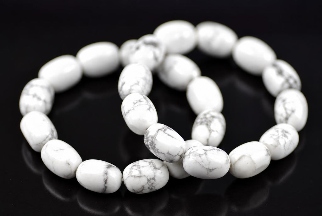 White oval howlite beads with grey veining arranged in overlapping bracelet loops on a dark reflective surface, viewed at an angle with softly blurred rear beads.