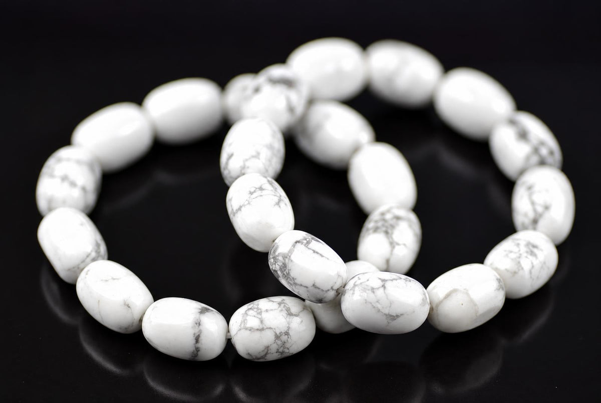 White oval howlite beads with grey veining arranged in overlapping bracelet loops on a dark reflective surface, viewed at an angle with softly blurred rear beads.
