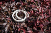 Two overlapping bracelets of white square beads with grey veining rest on dark burgundy foliage, viewed from above in sunlight.