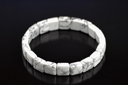 Howlite bracelet with white square beads and grey veining, arranged in a ring on a dark reflective surface, viewed at an angle.