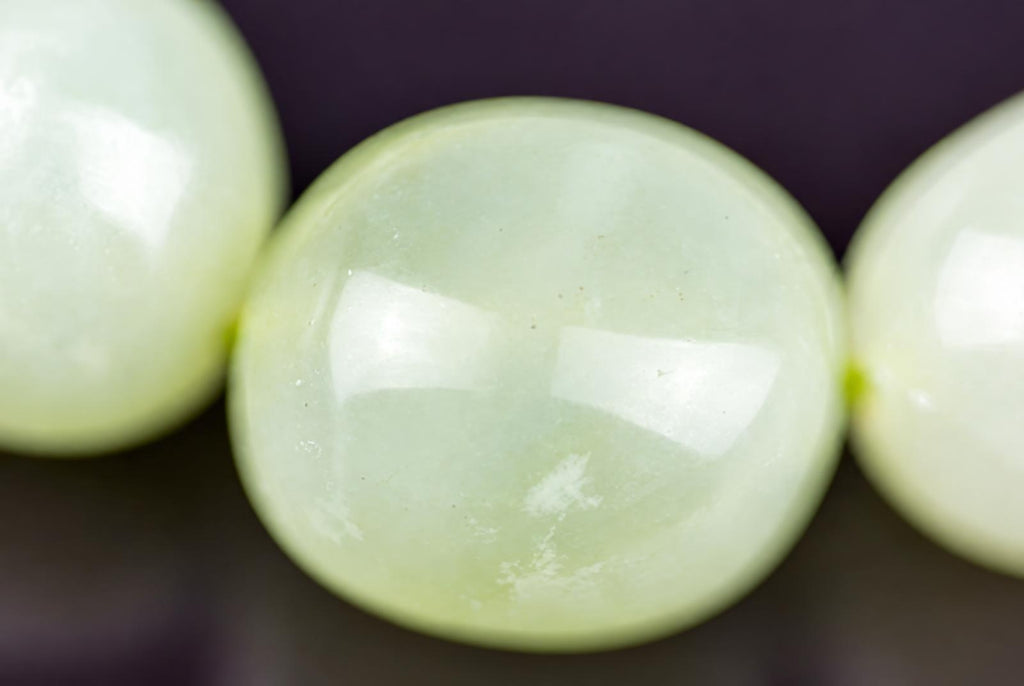 Close-up of pale green bracelet beads, with a glossy round central bead showing cloudy white markings and bright reflections, flanked by partially visible beads against a dark background.