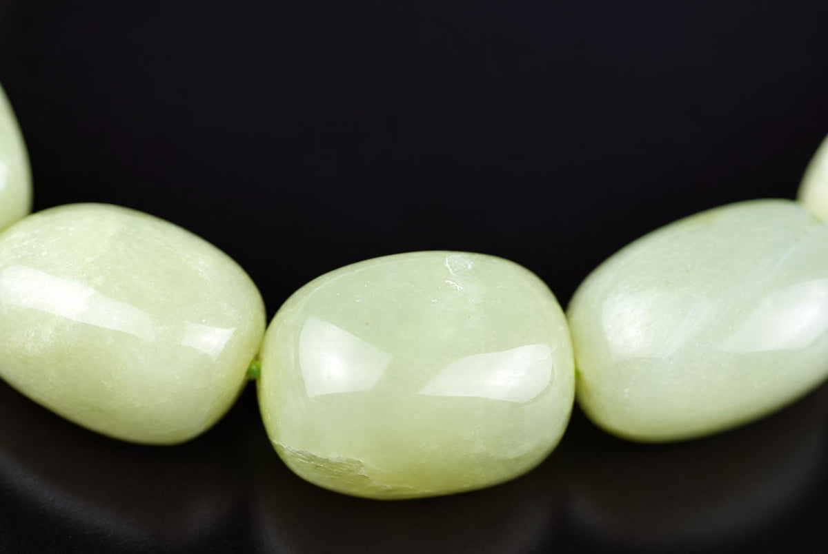 Close-up of polished pale green bracelet beads on a dark background, with a rounded rectangular bead between two oval beads and bright reflections on their surfaces.