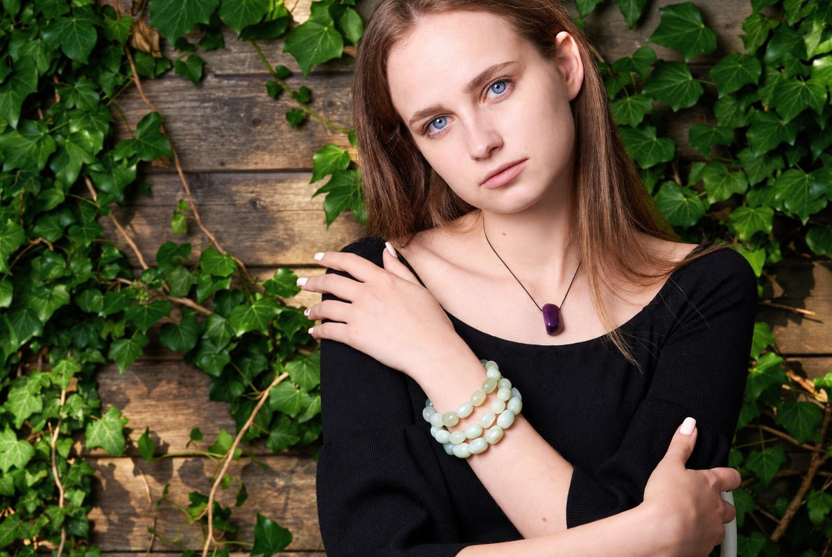 Pale green rounded-bead bracelet wrapped around a woman's wrist as she crosses her arms, wearing a black top and purple pendant against ivy-covered wooden boards.