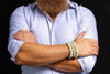 Pale green oval bead bracelets worn together on a wrist, shown across folded arms with a light blue rolled-sleeve shirt against a dark background.