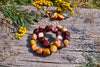 Two overlapping bracelets of polished, rounded beads in burgundy, mustard yellow, cream and pink on weathered wooden boards, surrounded by yellow flowers and greenery in sunlight.
