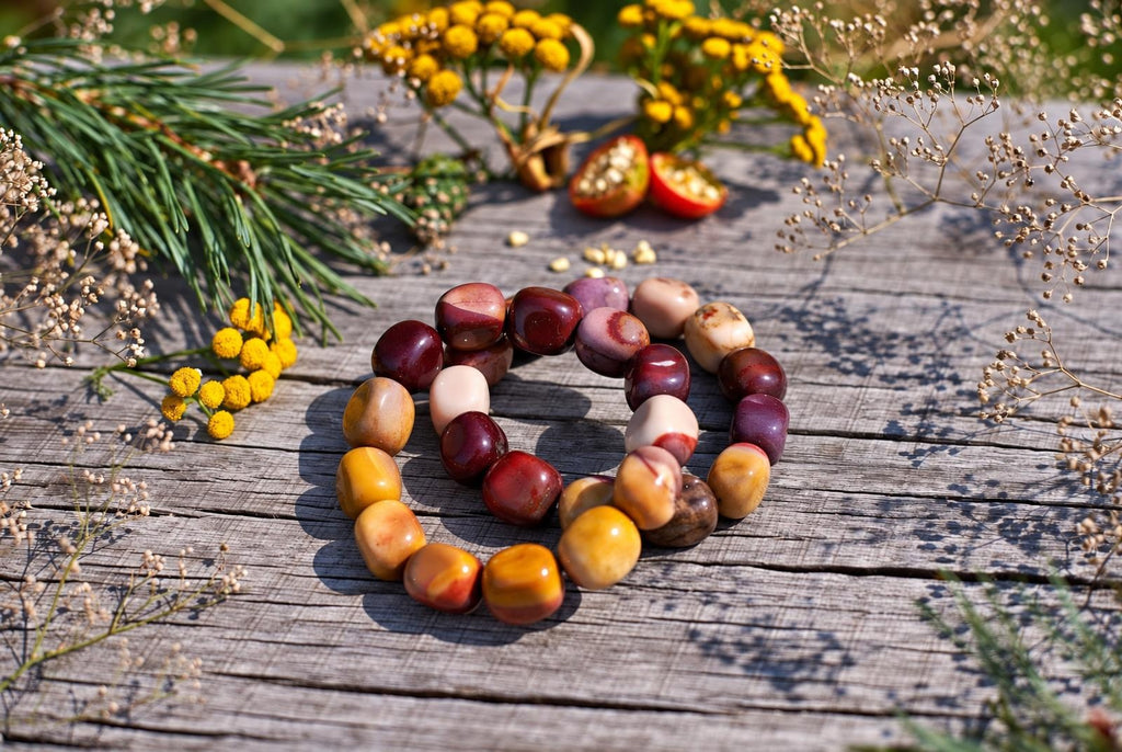 Two overlapping bracelets of polished, rounded beads in burgundy, mustard yellow, cream and pink on weathered wooden boards, surrounded by yellow flowers and greenery in sunlight.