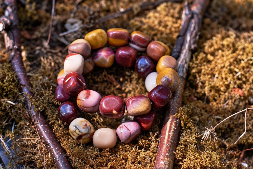 Overlapping loops of a polished, rounded bead bracelet in burgundy, mustard yellow, pink and cream, with varied natural markings, resting on brown moss among twigs in sunlight.