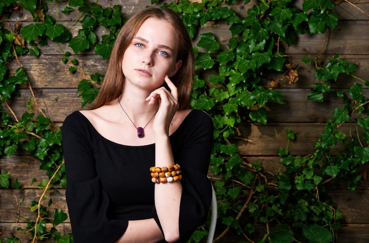 Three stacked bracelets with rounded yellow, ochre, cream and burgundy beads on a woman's raised wrist, worn with a black top and purple pendant against an ivy-covered wooden wall.