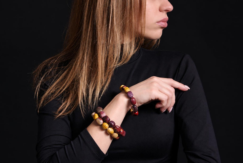 Glossy rounded-bead bracelets in mustard yellow, burgundy, pink and cream worn on a raised wrist, with black clothing and a dark background.