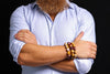 Three beaded bracelets in mustard yellow, cream and burgundy, with varied natural markings, worn on a wrist above crossed arms in a pale blue rolled-sleeve shirt against a dark background.