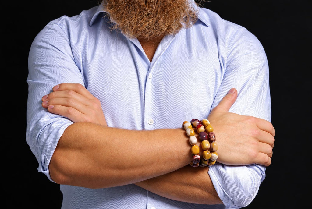 Three beaded bracelets in mustard yellow, cream and burgundy, with varied natural markings, worn on a wrist above crossed arms in a pale blue rolled-sleeve shirt against a dark background.