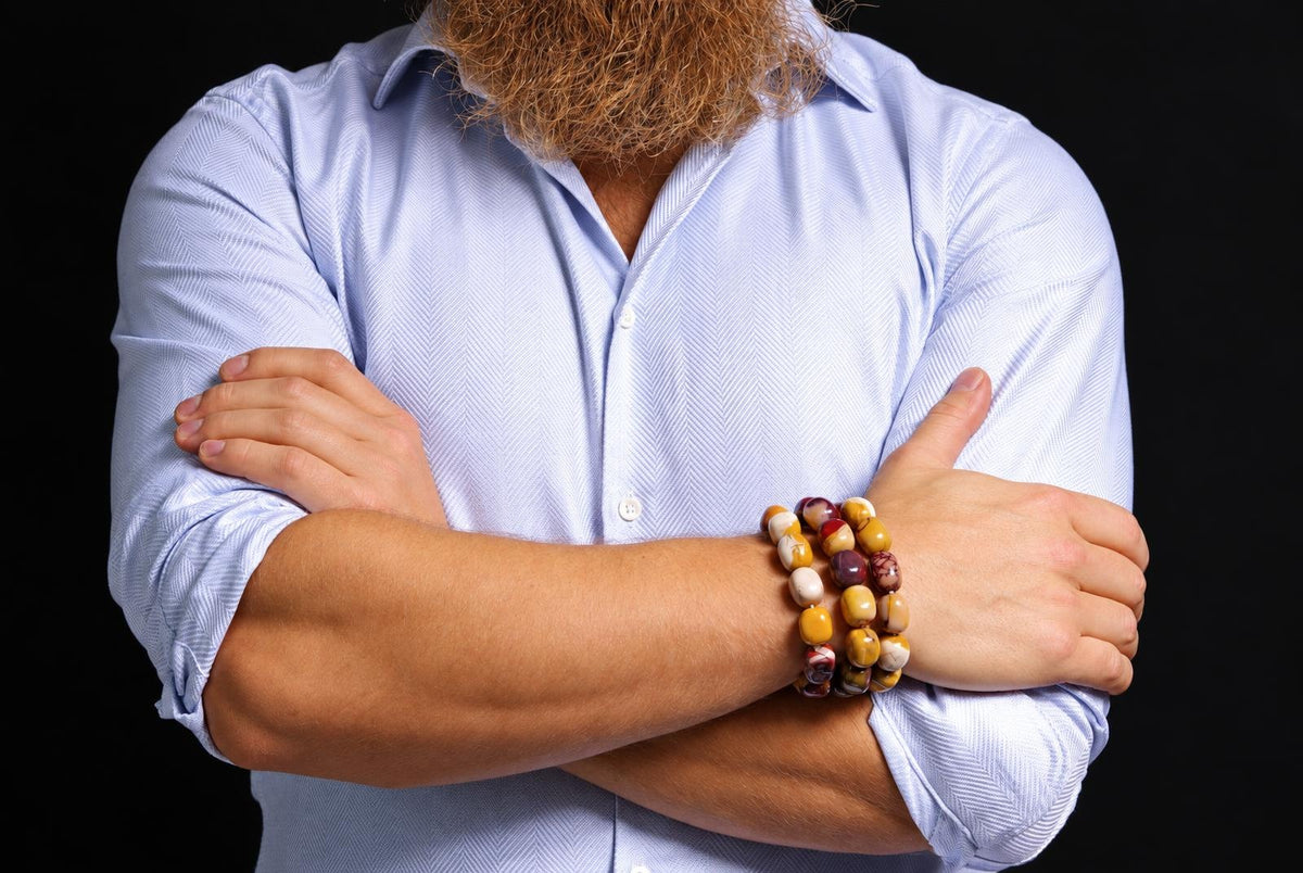 Three beaded bracelets in mustard yellow, cream and burgundy, with varied natural markings, worn on a wrist above crossed arms in a pale blue rolled-sleeve shirt against a dark background.