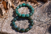Two overlapping bracelets of polished, rounded green and blue-green beads on coarse beige burlap beside tree bark, viewed from above in sunlight.