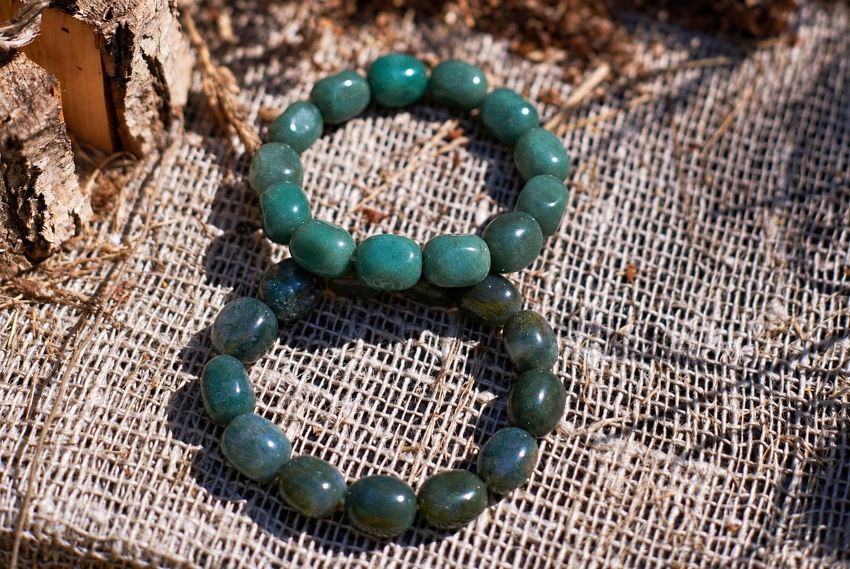 Two overlapping bracelets of polished, rounded green and blue-green beads on coarse beige burlap beside tree bark, viewed from above in sunlight.
