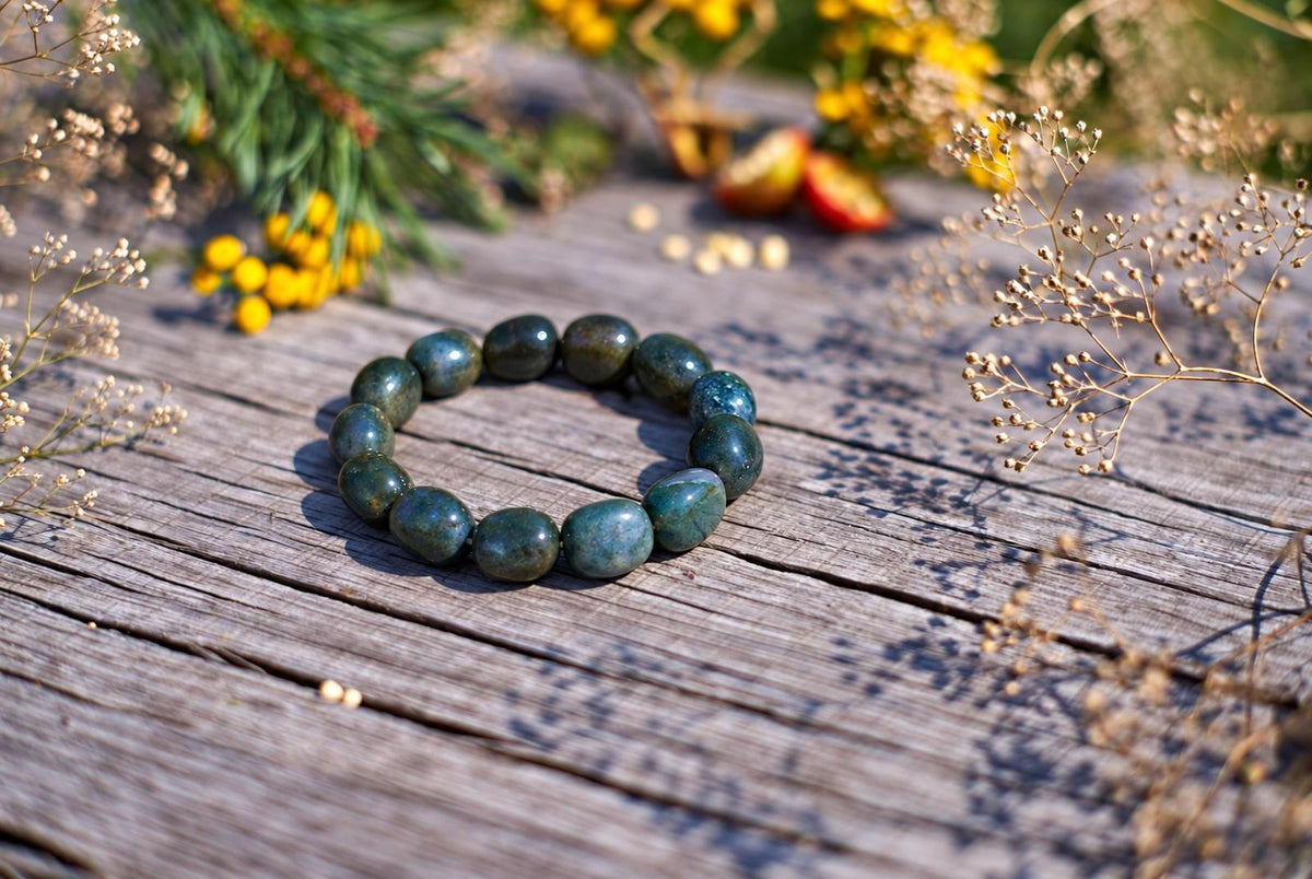 Glossy, rounded blue-green beads form a bracelet on weathered wooden boards, viewed at an angle in sunlight, with yellow flowers and delicate dried stems around it.