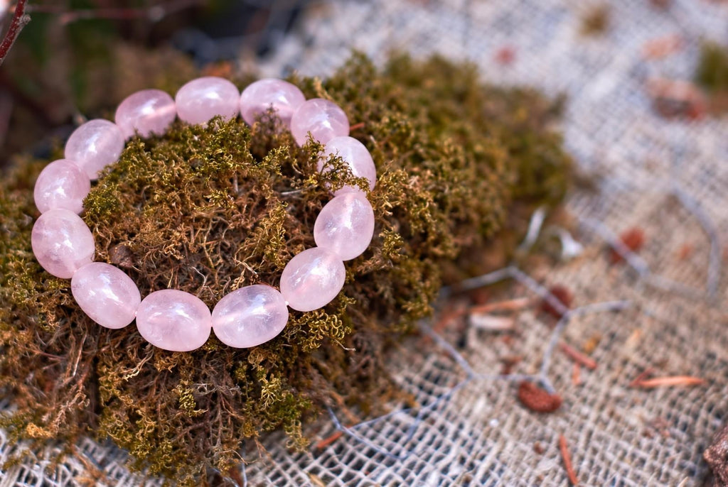 Pale pink bracelet with polished, translucent oval beads arranged in a loop on brown-green moss, viewed close up at an angle with pale mesh in the background.