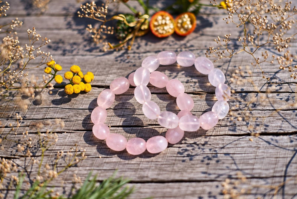 Two overlapping bracelets of polished, translucent pale pink oval beads on weathered wooden boards, surrounded by yellow flowers and delicate dried stems in sunlight.